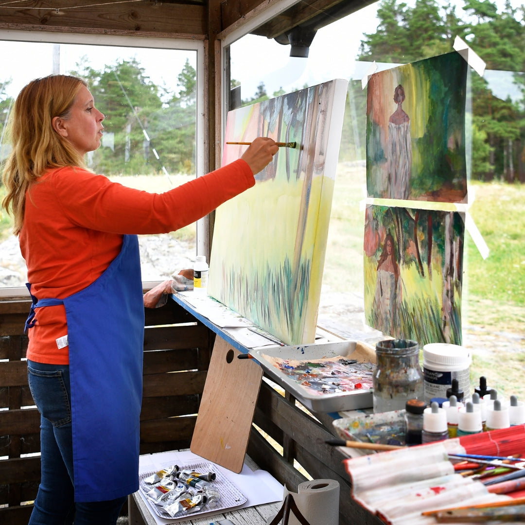 Artist Ingela Johansson painting on a canvas outside in a red sweater and blue appron